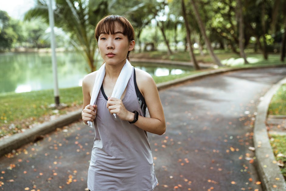 Serious young ethnic Asian female wearing sport clothes and smart watch running along pond in park and looking away while holding towel